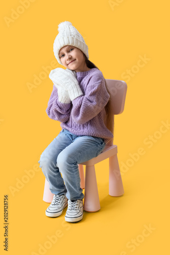 Cute little girl in warm clothes sitting in chair on yellow background