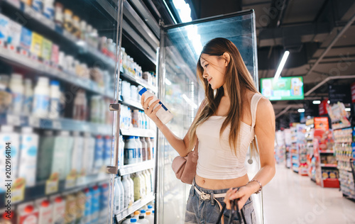 Young asian woman customer supermarket shopping cart choosing dairy products beverages shelf grocery food at department store