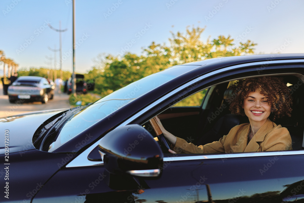 Fototapeta premium A smiling woman sits behind the wheel of a sleek car, looking out from the window with a carefree lifestyle vibe, enhanced by retro film color and warm, sunny tones.