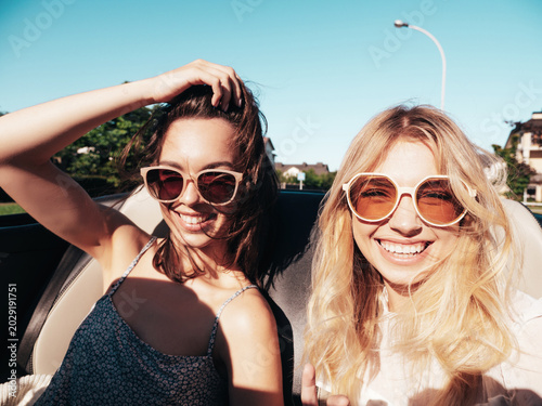 Portrait of two young beautiful and smiling hipster female in convertible car. Sexy carefree women driving. Positive models riding and having fun outdoors. Enjoying summer days. Cheerful and happy