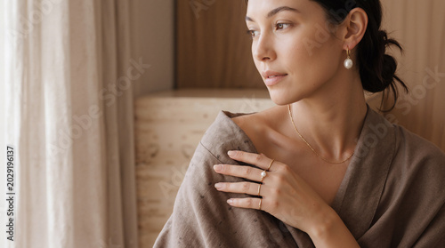 Elegant woman wearing pearl jewelry and soft robe by window in warm natural light