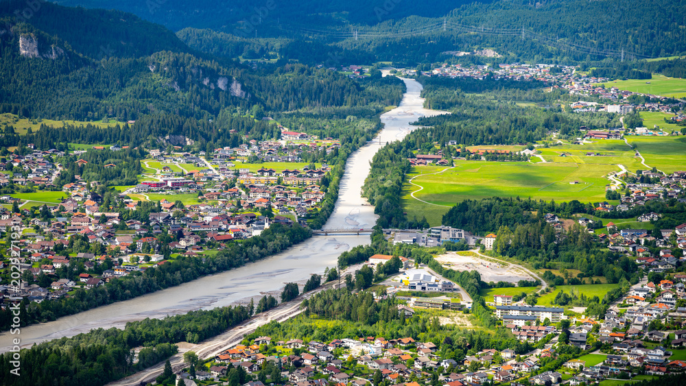 Fototapeta premium Aerial view shows the river flowing through the town of Reutte. Houses line the riverbanks, and green fields spread across the landscape. Trees and mountains surround the area.