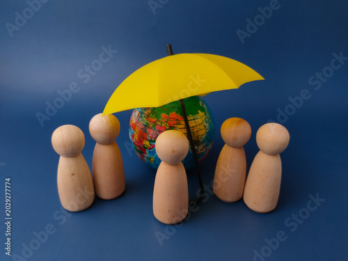 Five wooden peg dolls stand around a small globe sheltered by a miniature yellow umbrella on blue background