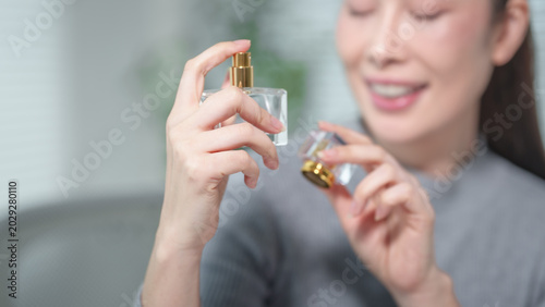 Close-up of smiling woman spraying perfume from glass bottle with gold atomizer, soft focus indoor background, beauty and fragrance concept.