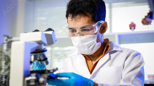 Male scientist wearing mask, safety goggles and blue gloves using microscope in modern laboratory, close-up with copy space.