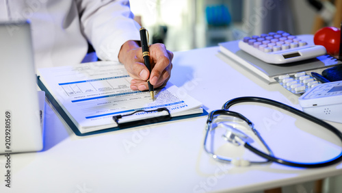 Obraz na plátně Doctor’s hand filling out medical form on clipboard at clinic desk with stethosc
