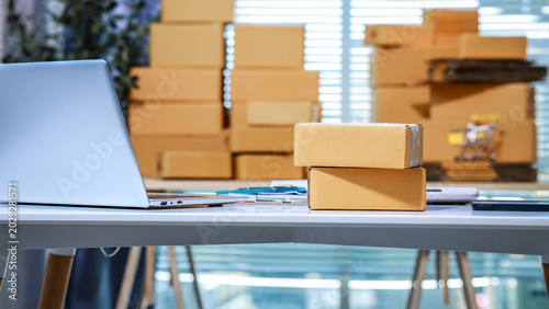 Office desk with laptop and stacked cardboard boxes, blurred boxes in background, daylight through blinds; ecommerce shipping, small business logistics and packing concept.