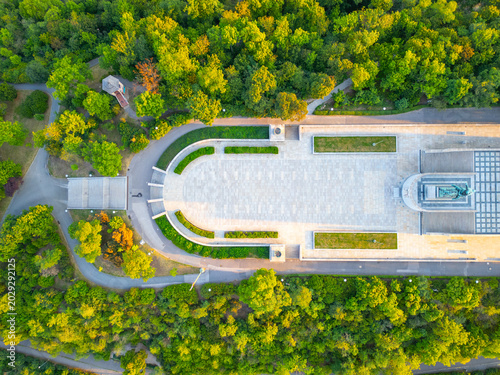 The National Monument at Vitkov is seen at sunrise. The area is surrounded by trees, and the Tomb of the Unknown Soldier is visible. People can be seen walking and enjoying the morning.