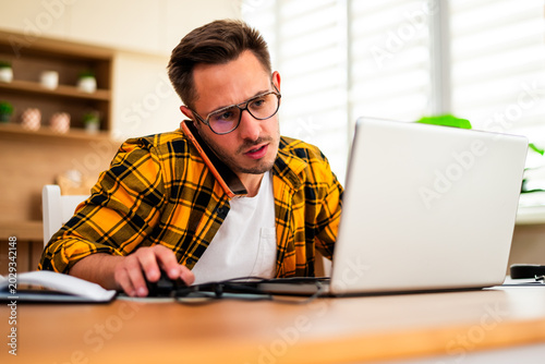 Young man is working from his home. He is using laptop and talking on smartphone. He appears worried.