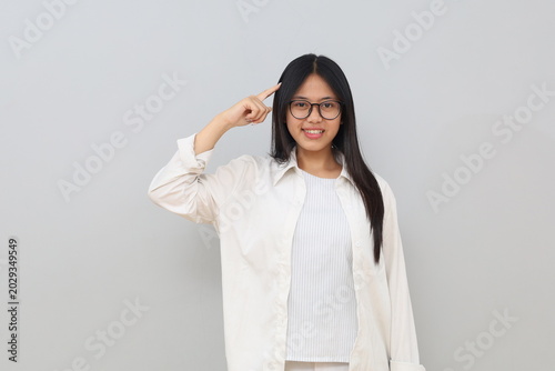 A young Asian woman looking confused or making a mistake while scratching her head, wearing casual clothes and glasses, looking at the camera, standing isolated on a white background.