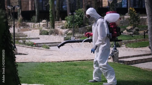 Worker in protective suit spraying garden with pesticide using backpack fogger, outdoor pest control treatment in residential yard