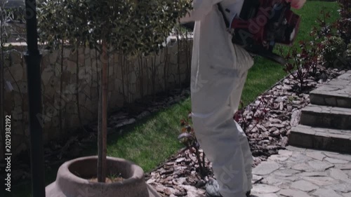 Worker in protective suit spraying garden with pesticide using backpack fogger, outdoor pest control treatment in residential yard