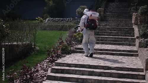 Worker in protective suit spraying garden with pesticide using backpack fogger, outdoor pest control treatment in residential yard