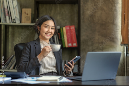 Professional Asian Businesswoman Smiling at Desk with Laptop, Smartphone, and Coffee Cup in Modern Office.