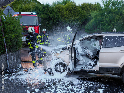 Firefighters extinguishing burning passenger car with foam during emergency response operation on residential street
