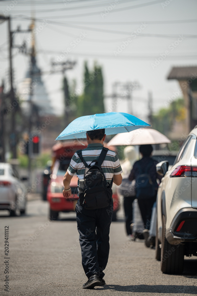 Fototapeta premium Man walking through city street holding umbrella, health safety and protection from heatstroke during heatwave, male hiding from sun under umbrella while passing by crossroad on hot summer day.