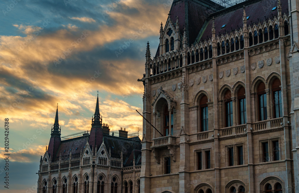 Obraz premium Majestic view of the Hungarian Parliament Building in Budapest at sunset with dramatic golden clouds over Gothic Revival architecture and historic stone facades