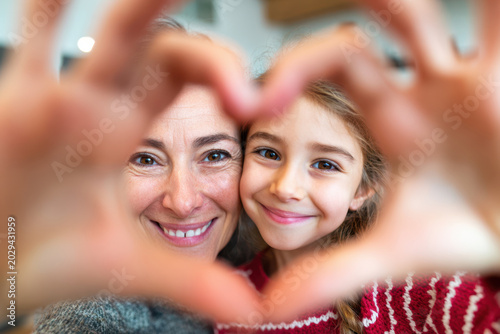 Mother and daughter form heart shape with hands, smiling together in a cozy indoor setting