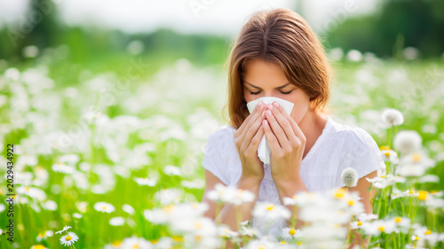 Young woman sneezing into tissue in daisy field with spring allergy