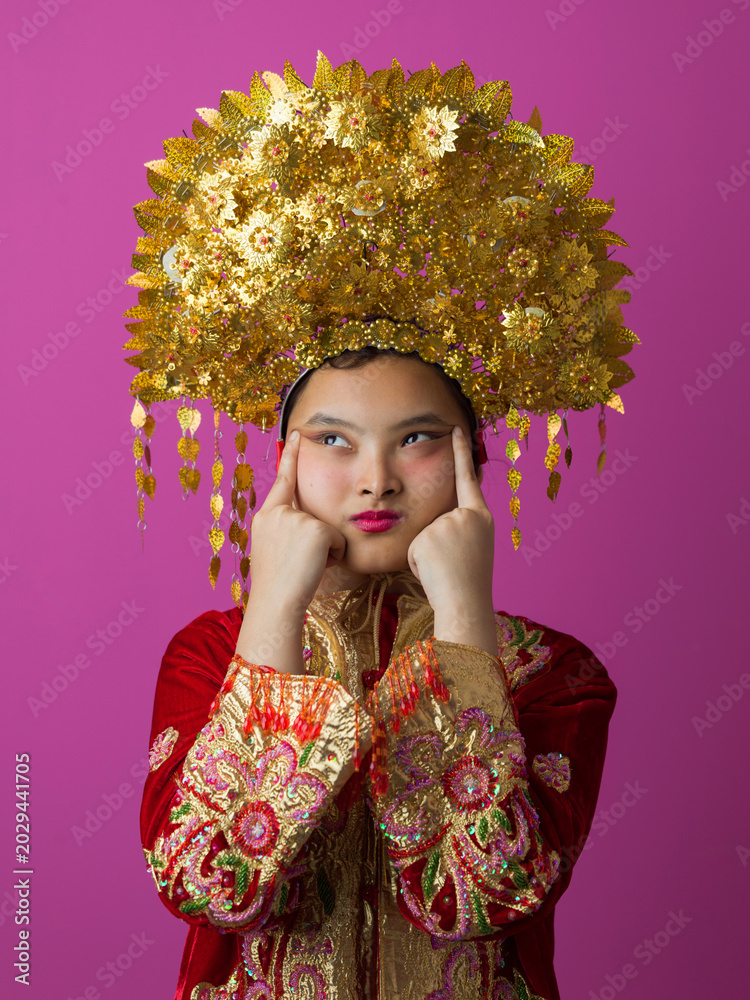 Fototapeta premium Young Indonesian Asian Girl Wearing Ornate Gold Headdress and Embroidered Traditional Costume Against Pink Background