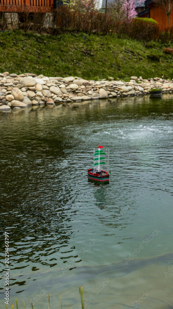 Obraz premium Small toy sailboat with green sails navigates a tranquil pond with a fountain