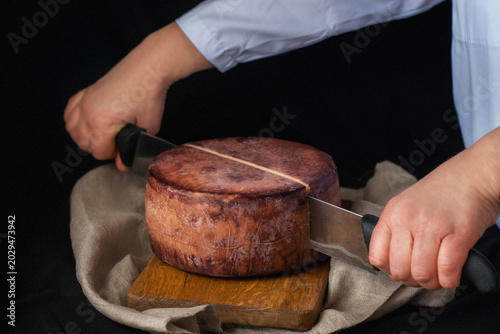 A cheesemaker cuts a wheel of hard cheese with a knife. Exquisite cheese in wine on a wooden board. A farm product made from milk on a dark background. Delicious hard cheese. Wheel of cheese. Perfect 