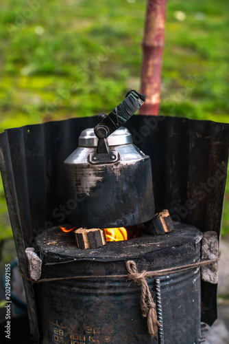Quadro em tela A rustic metal kettle heating on a traditional charcoal stove outdoors, surround
