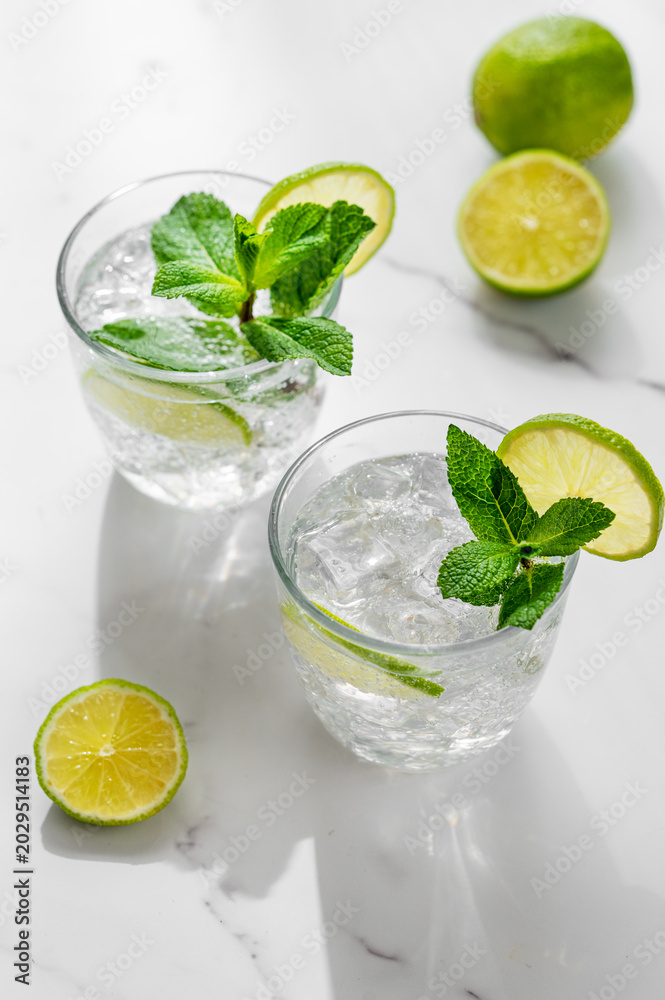 Fototapeta premium A fresh mojito with lime, mint and ice in glasses. Citrus lemonade or cocktail on a marble table with shadows.
