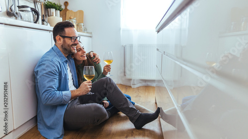 Obraz na płótnie Happy Couple Sitting on Kitchen Floor Drinking Wine and Embracing in Cozy Home M