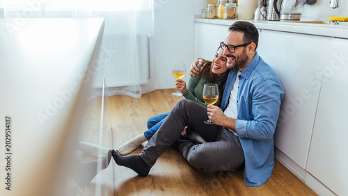 Fotografie Happy Couple Sitting On Kitchen Floor Drinking Wine Together During Cozy At-Home