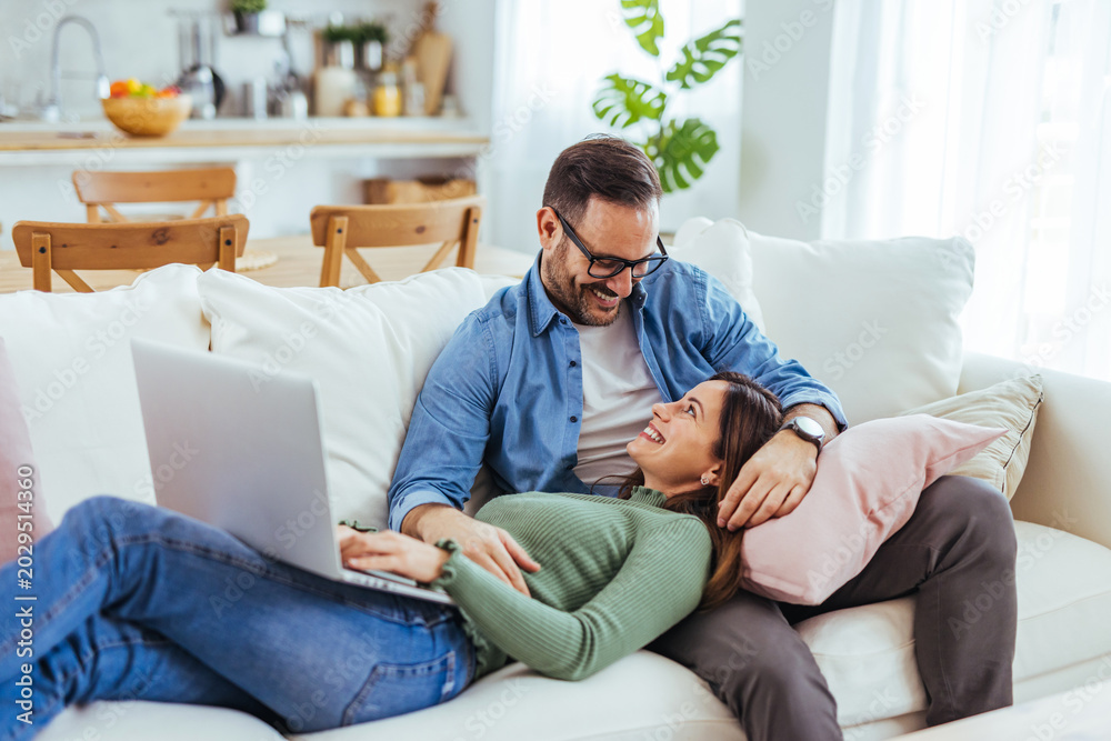 Obraz premium Smiling Couple Relaxing on Sofa at Home Using Laptop Together in Bright Living Room