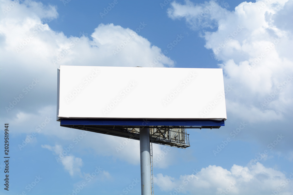 Fototapeta premium Blank white billboard rises over open blue sky with soft clouds and leaves clean copy space for outdoor advertising mockup.