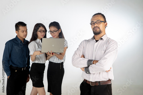 Confident Leader Standing with Arms Crossed While Team Works on Laptop in Background