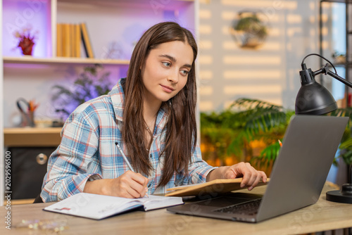 Schilderij op canvas Young woman on home table studying with laptop and book while writing notes for online education at home
