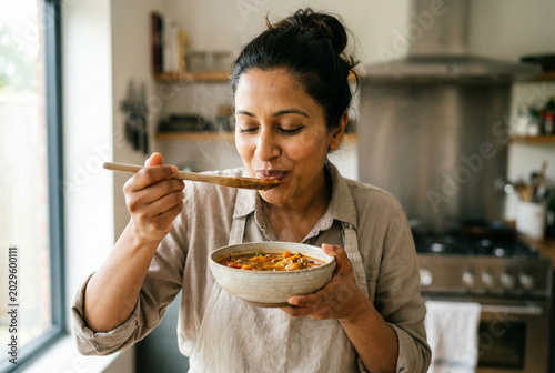 Indian woman savoring aroma of homemade vegetable soup in bright modern kitchen wearing apron with wooden spoon