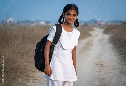 South Asian girl student white school uniform carrying black backpack standing on rural dirt road morning light