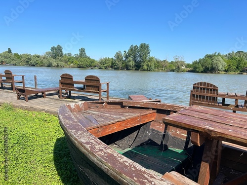 Foto Old Wooden Boat on River Pier with Bench Chairs and Blue Sky
Abandoned rustic wooden boat on riverside pier with wooden bench chairs overlooking calm river and green trees