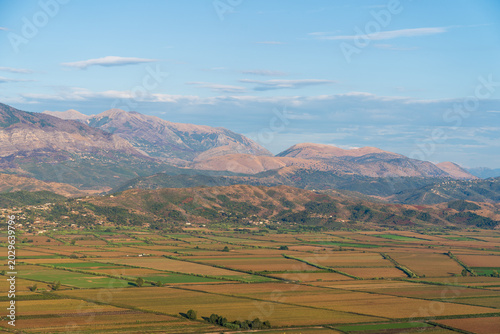 Saranda, Albania - August 26, 2025: Panoramic view of agricultural green fields in the valley surrounded by high mountain ranges under a blue sky on a sunny evening