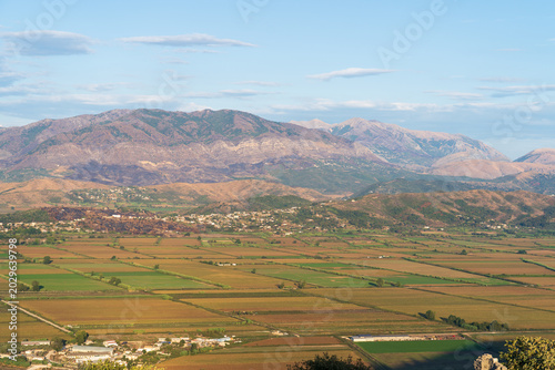 Saranda, Albania - August 26, 2025: Panoramic view of agricultural green fields in the valley surrounded by high mountain ranges under a blue sky on a sunny evening