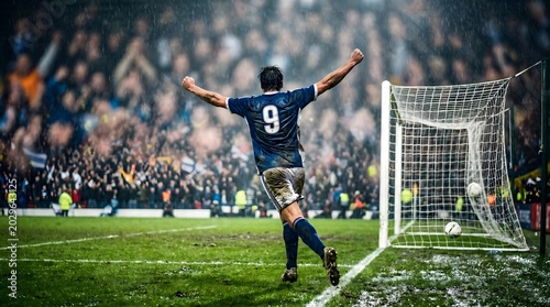 A male soccer player in a blue jersey with number 9 celebrates scoring a goal on a muddy field in front of cheering fans.