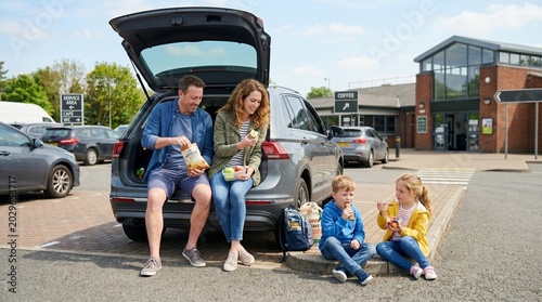 Family having a picnic break at a service station, with car and building in background