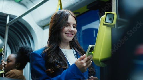 Pretty Caucasian woman with long hair holding smartphone and validating ticket on public transport. Female smiling while using contactless payment system inside modern city bus at night.