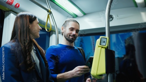 Man in blue sweater holding smartphone to yellow payment terminal smiling while standing in tram. Woman beside him looking pleased. Friends using mobile technology during ride on public transport.