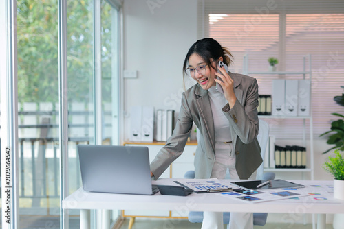 Asian businesswoman working on laptop, talking on smartphone, and multitasking at a modern office desk, smiling confidently