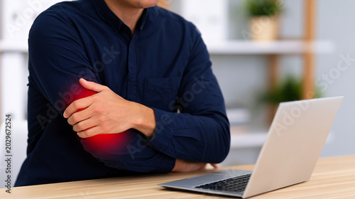 Elbow Pain. Unrecognizable worker gripping elbow with red highlight beside desk and laptop, blurred office background. Concept of chronic tennis elbow pain and repetitive strain injury.