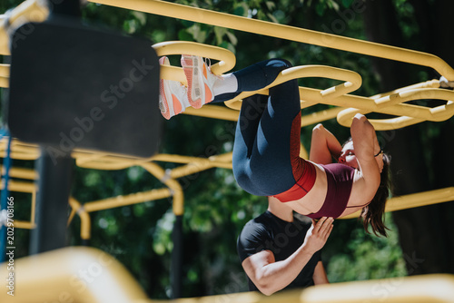 Canvas Print A woman performs hanging leg raises on yellow outdoor monkey bars with a trainer assisting
