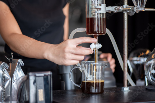 Photos Smiling Asian woman adjusting a valve on a glass coffee drip apparatus to brew craft coffee in a workshop setting