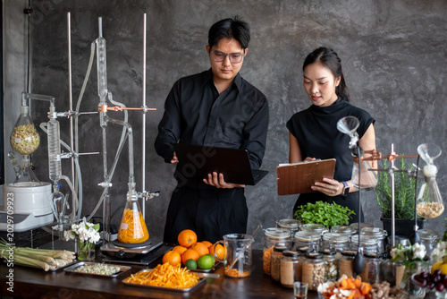 Foto Smiling Asian man and woman in black outfits discussing research with a laptop and clipboard near a distillation apparatus in a laboratory