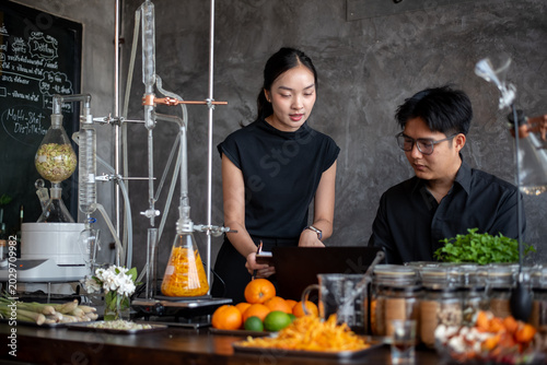 Quadro em tela Smiling Asian man and woman in black outfits discussing research with a laptop and clipboard near a distillation apparatus in a laboratory
