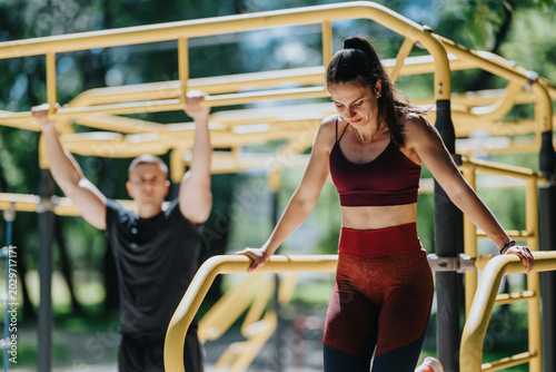 A fit woman performs calisthenics on yellow outdoor bars while a partner trains in the background. The scene shows an outdoor workout in a park with strength training equipment and focused athletes.